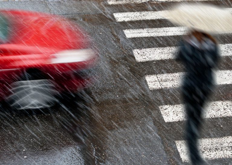 A person holding an umbrella, crossing a pedestrian crossing in the rain, as a red car approaches.
